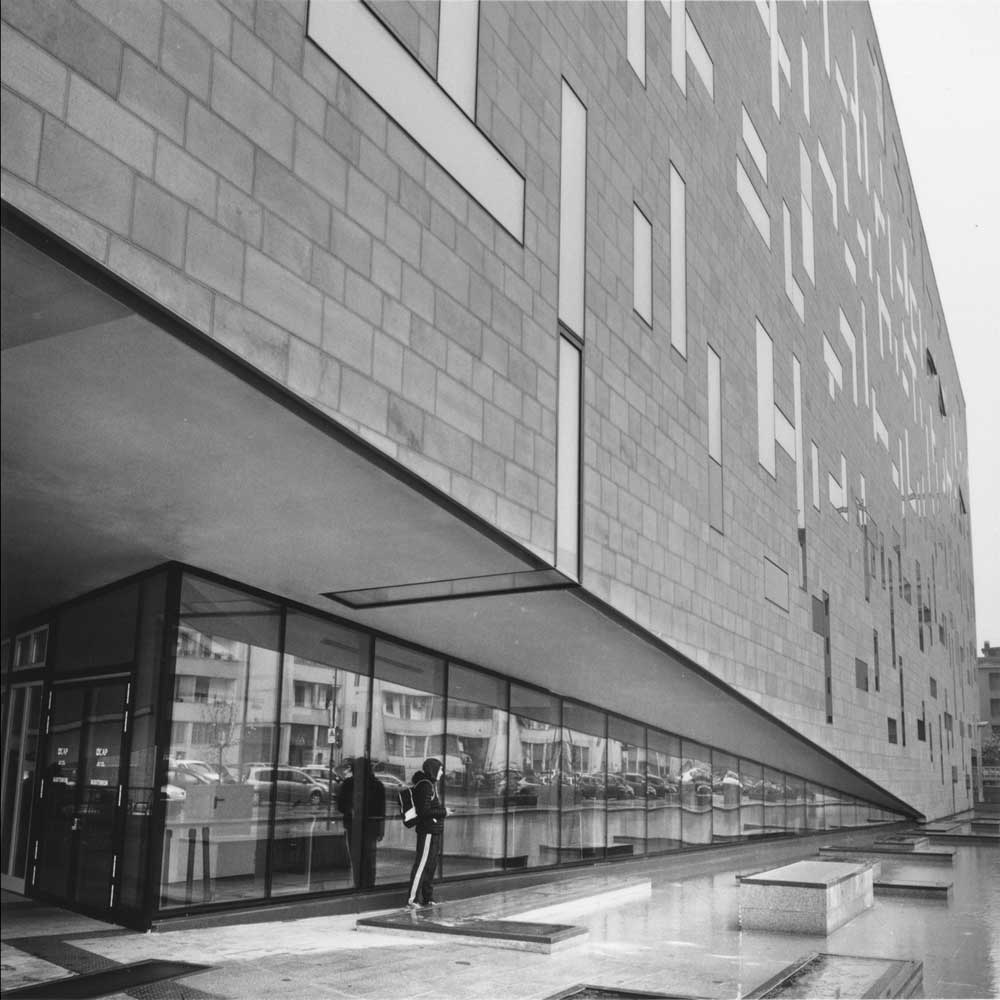 Una persona medita sotto il cornicione di un edificio moderno riflettendosi nelle vetrine e nelle fontane